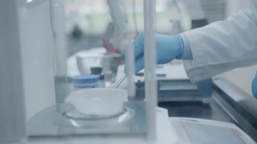 Person wearing lab coat and gloves spoons white powder into a small bowl on a scale