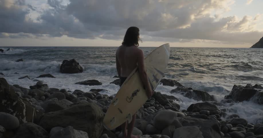A surfer stands on a rocky beach at sunset