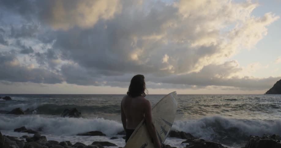 A man stands on a beach with a surfboard at sunset