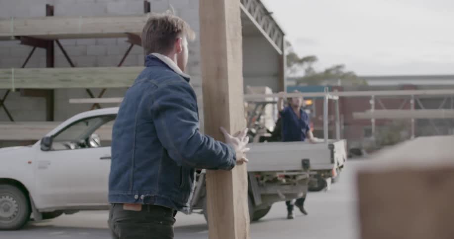 A man is picking up a wood board and carrying it toward a truck