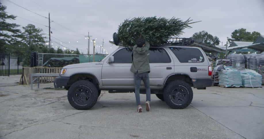 Man carries a pine tree to a gray truck and puts the tree on the roof