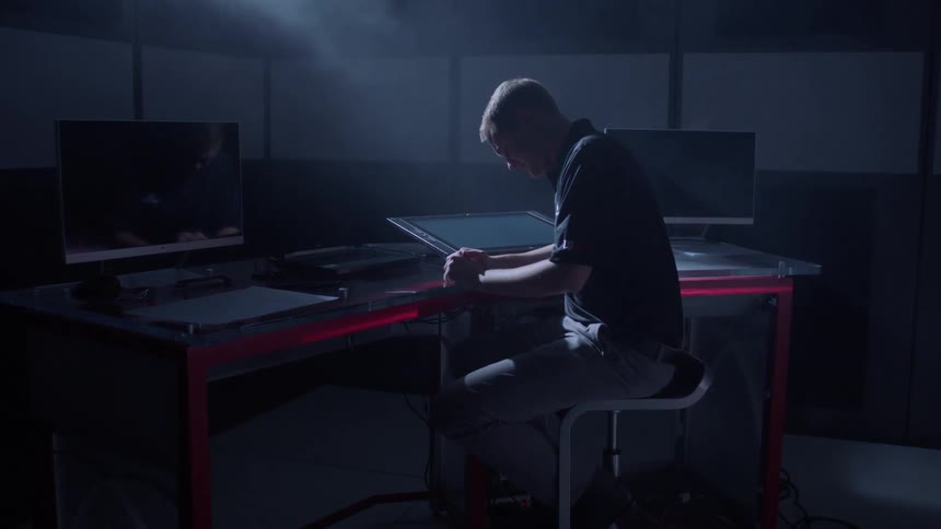 A man sits with head bowed at a workstation with several monitors