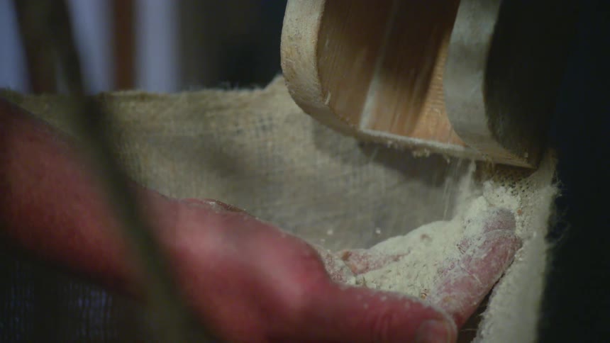 A hand catches ground grains from a chute
