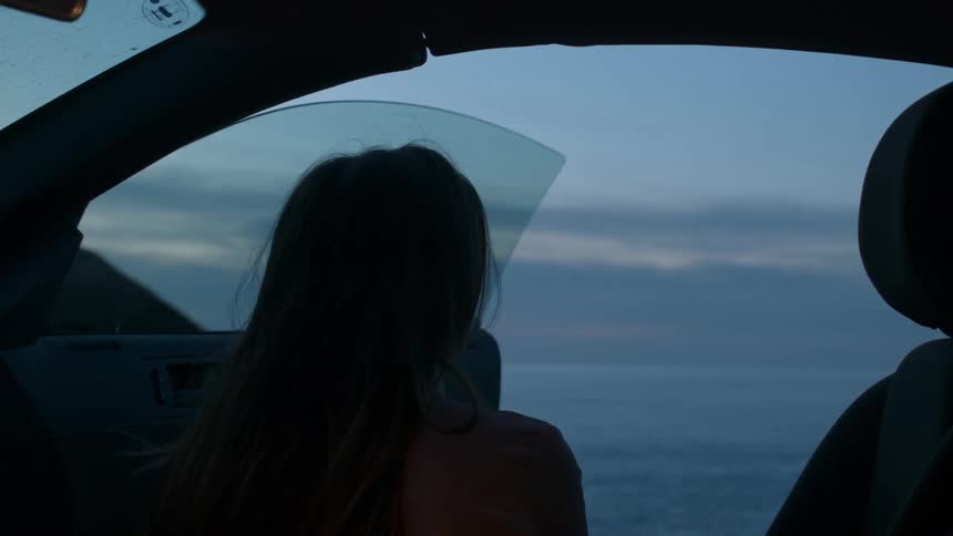 A woman sits in the passenger seat of a car on the coast