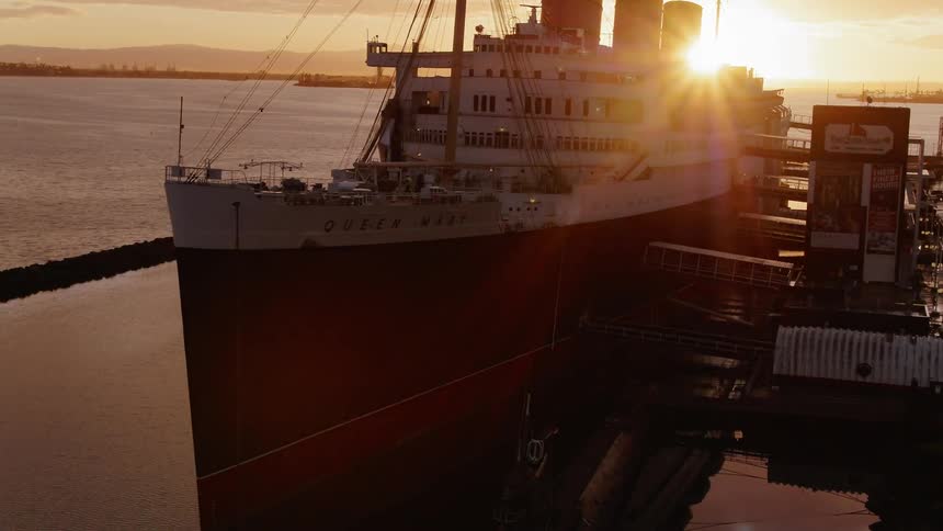 Birds are flying in the air near a ship docked in a harbor at dusk