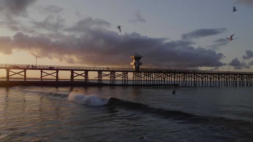 Surfers sit on their boards near the pier at sunset