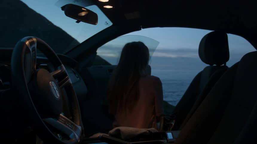 A woman is sitting in the passenger side of a car looking out the door at the ocean