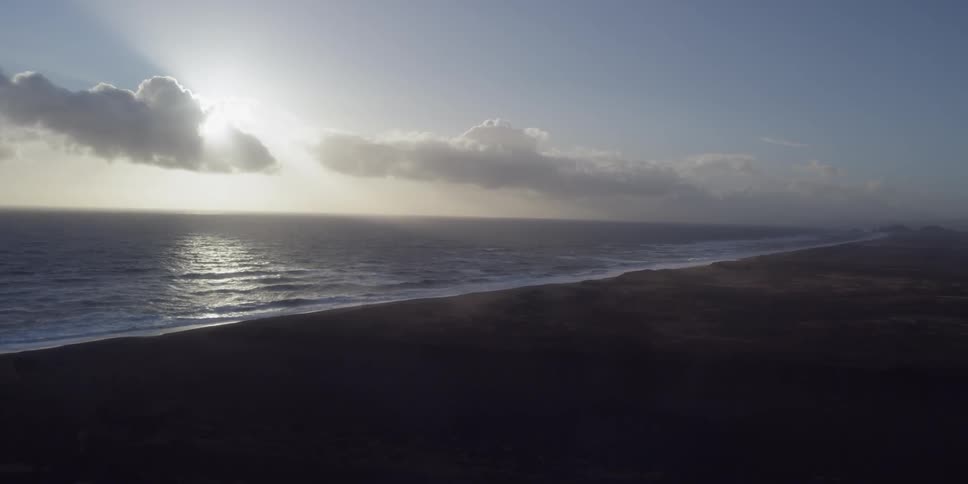 The sun is peeking through clouds as waves roll onto a beach