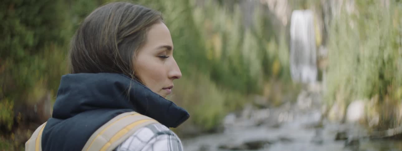 A woman in a jacket is standing in the woods looking at a large waterfall