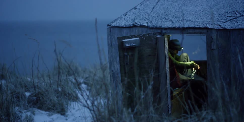 Person puts socks on in a small wooden shack on a snowy evening