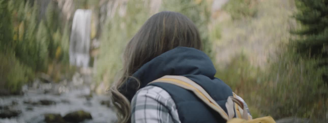 A woman wearing a backpack walks over rocks toward a waterfall