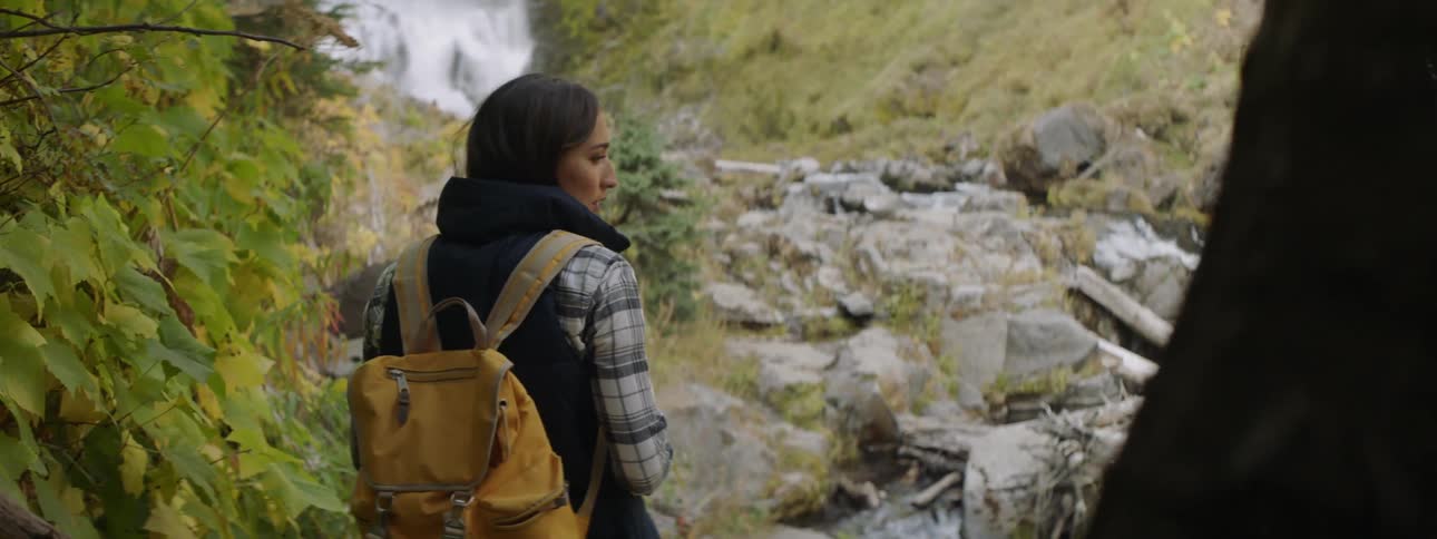 A woman wearing a backpack looks around out by the stream