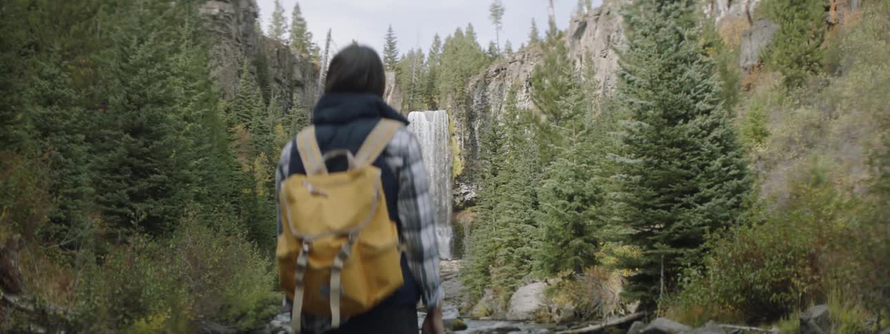 Woman wearing a yellow backpack stands in the woods near a waterfall