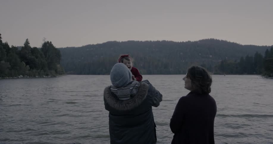 Couple and their infant next to a lake on an overcast day as the man lifts the baby into the air
