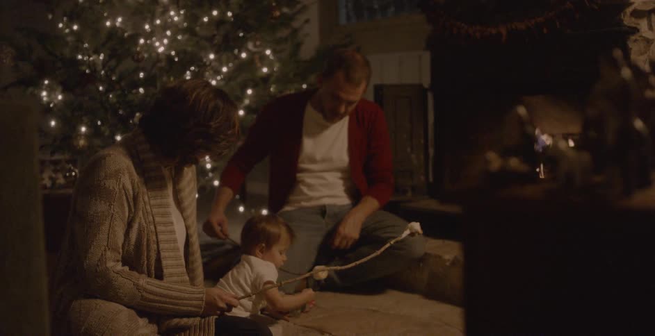 Man and woman sit with child in front of a fire indoors roasting marshmallows
