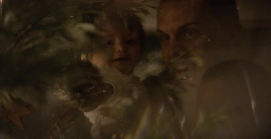 A dad holds his daughter as she reaches for an ornament on the tree