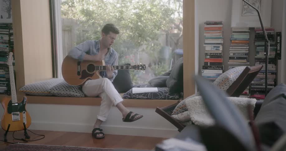 A man is sitting in a window playing a guitar and writing a song during the day
