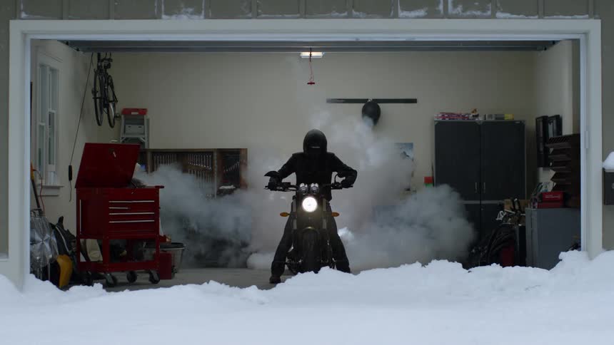 Someone is sitting on a motorcycle while there is a lot of snow on the ground outside the garage