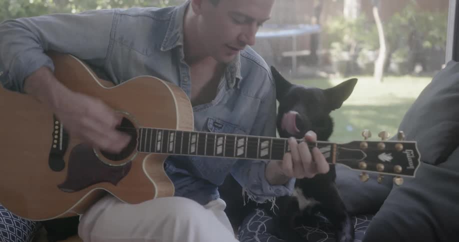 A man is strumming a guitar while his dog sits next to him inside during the day