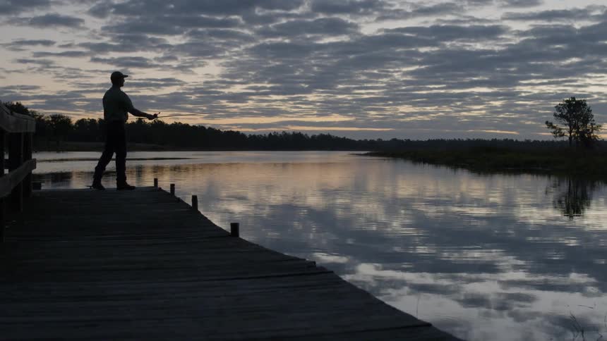 Man casting a line into the waters while standing on a dock on a cloudy day at sunrise