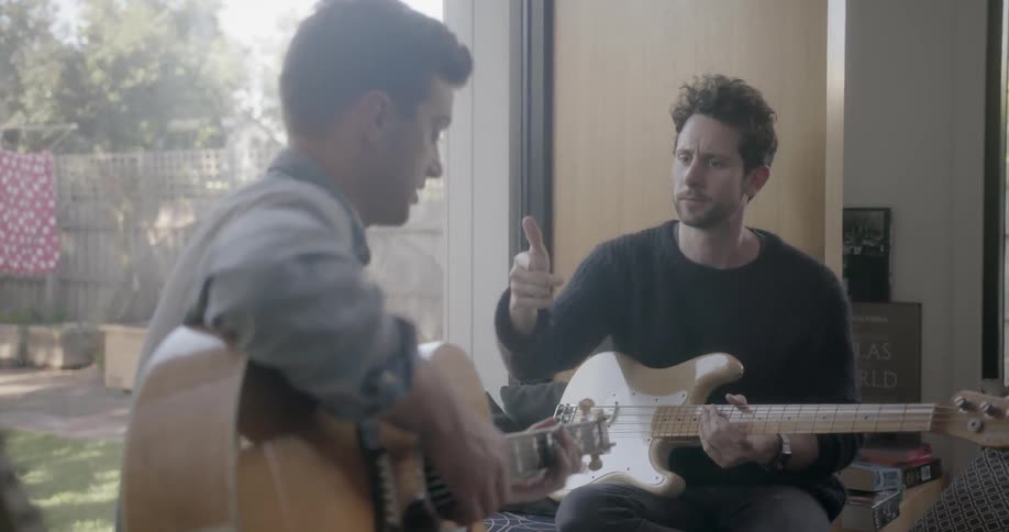 A man is giving advice to another man as they play guitars inside during the day
