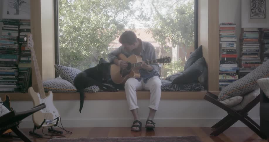 A man sits on a window seat playing a guitar and his dog jumps down