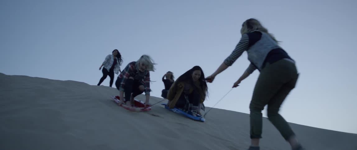 A group of friends are sand boarding down a sand dune at dusk