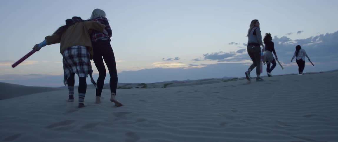 Women run up a sandy hillside in a desert at sunset