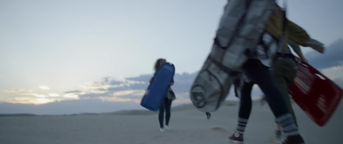 A group of friends are carrying sand boards as they walk over a sand dune at dusk