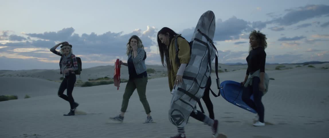 A group of friends are carrying sand boards as they walk over a sand dune at dusk