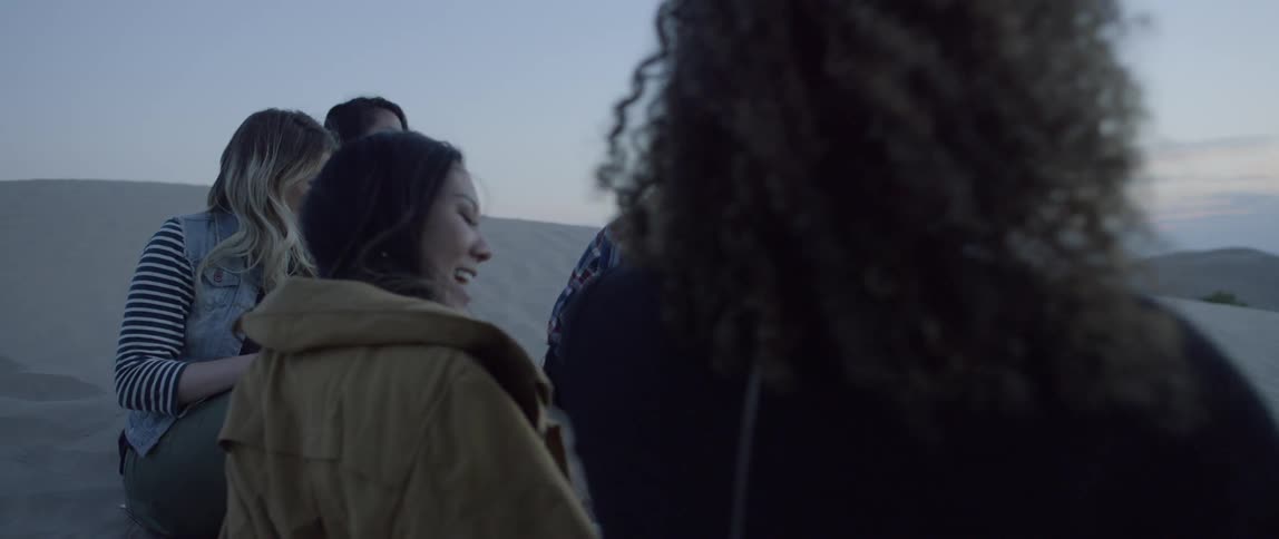 A group of female friends is sitting on a sand dune and socializing at dusk