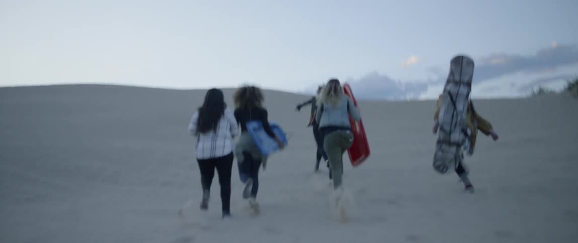 Group of women run across sandy hills carrying sleds