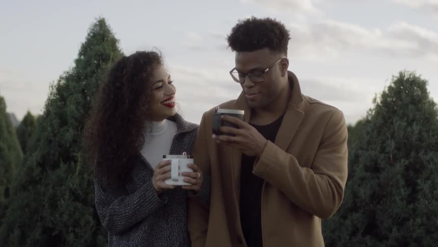 A man and woman are drinking from mugs as they stand in a christmas tree farm during the day