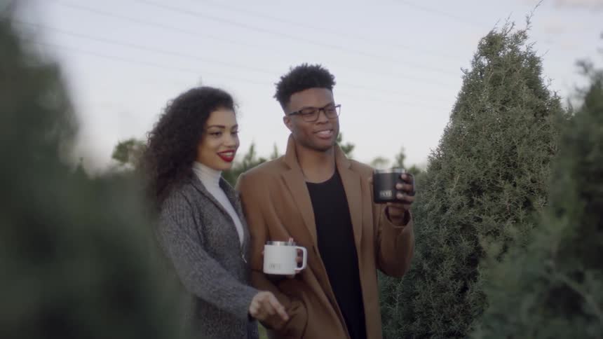 Couple walking and looking at bare christmas trees while they carry their coffee mugs