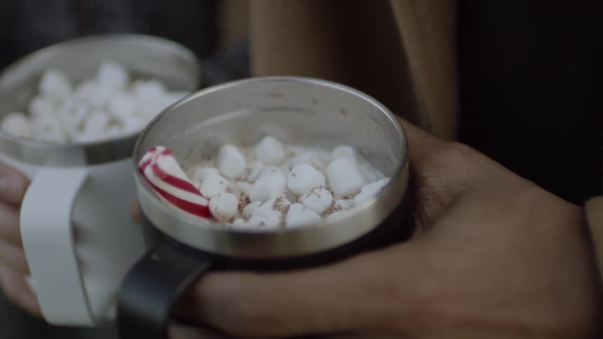 A couple holds mugs of hot chocolate with marshmallows and candy cane on top