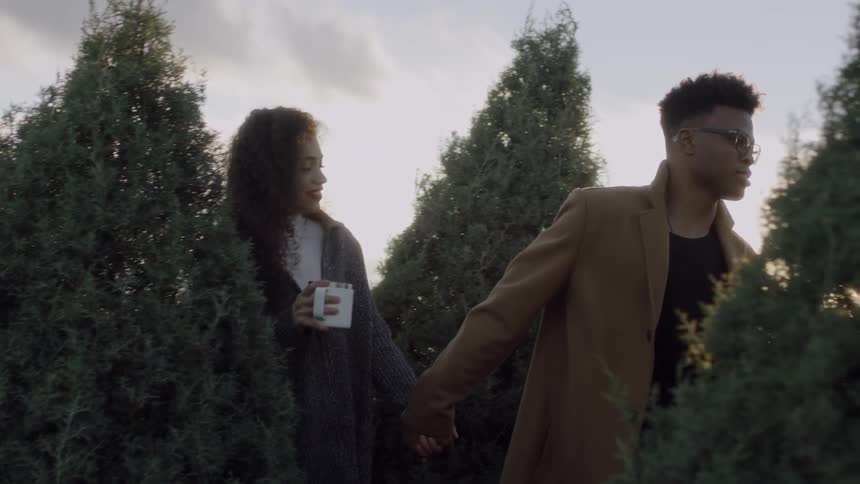Couple walking and looking at bare christmas trees while they carry their coffee mugs