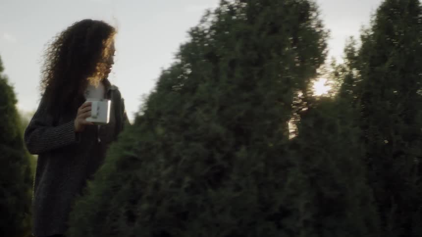 A man and woman are walking through a christmas tree farm holding hands and mugs