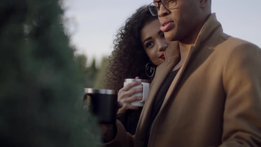 A man and woman inspect a tree while holding coffee mugs