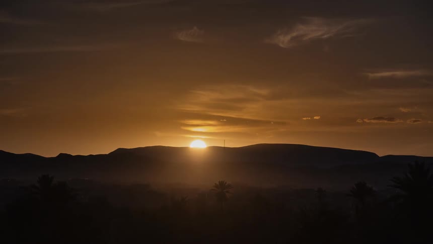 The sun is setting beyond hills in a desert on a dusty day