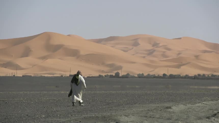 Person walking across a barren area using a walking staff with a town in the distance