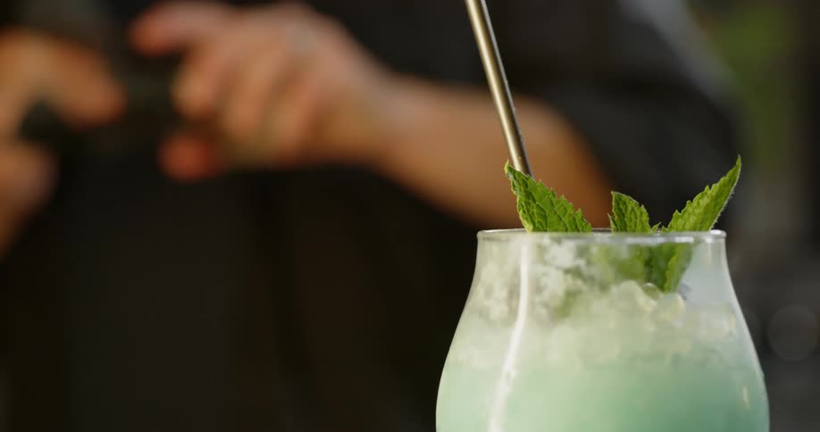 A bartender cleans a glass while a straw sticks out of a cocktail with mint leaves