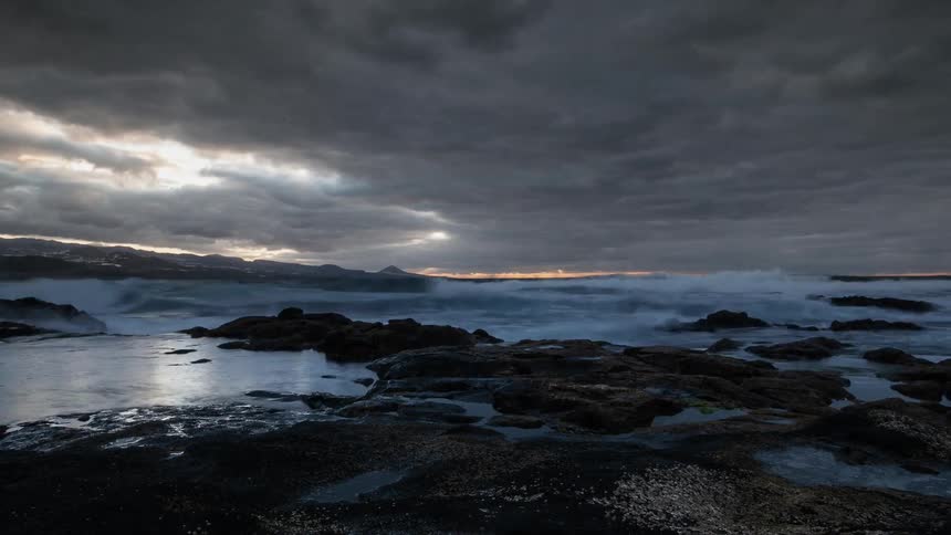 Clouds are moving over a rocky beach and coastline at dusk