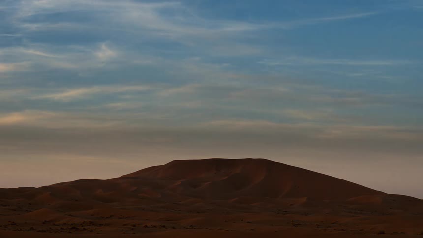 Clouds are drifting in the sky above a desert as day turns to night