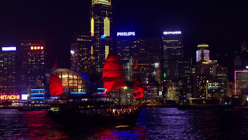 A chinese junk sails through a harbor in a metropolitan area at night