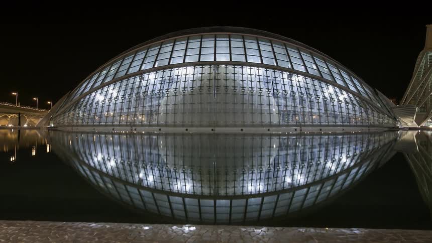 A building and bridge are reflected on the water's surface at night