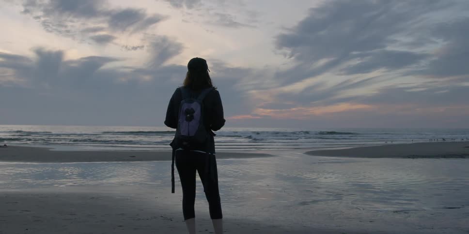 A woman wearing a backpack stands on the sand and looks at the ocean