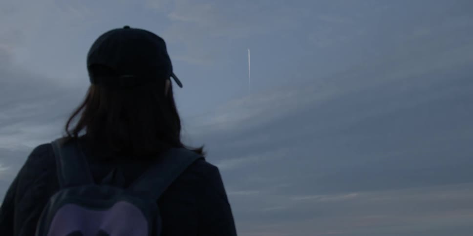 A woman is watching a rocket ascend in the sky at dusk