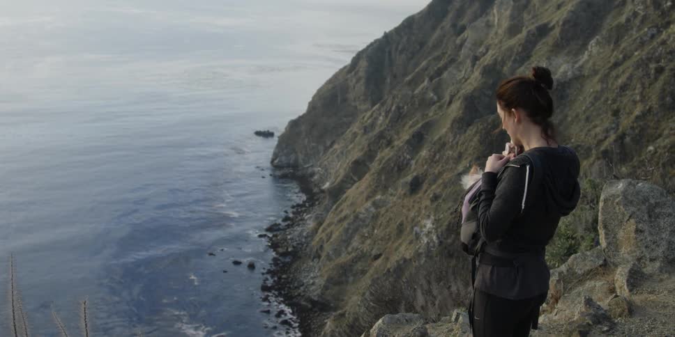 A woman stands on a coastal cliff with a dog strapped to her chest