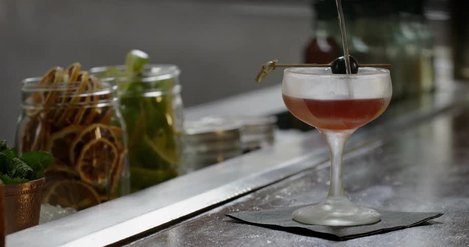 Pitcher pours liquid into a glass on a table surrounded by jars filled with various foods