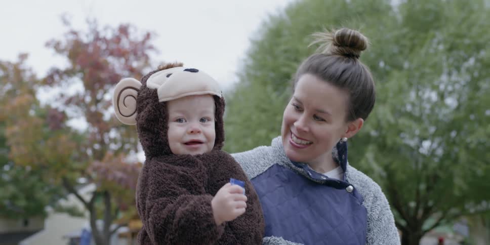 A mother is smiling as she holds her toddler and walks outside during the day
